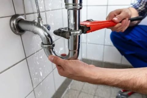 A plumber is using a large red wrench to repair a chrome P-trap pipe under a sink, with water leaking and spraying from the connection point.